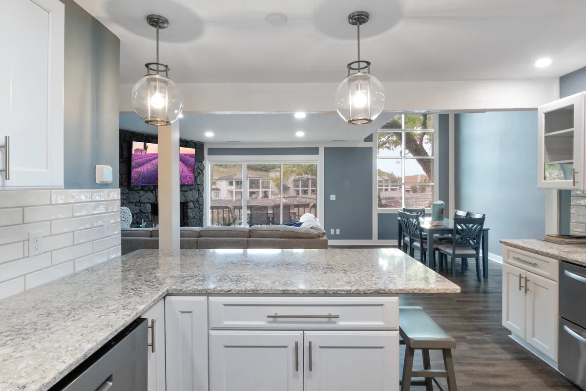 An open kitchen layout showcasing a granite countertop, pendant lighting, large windows flooding the space with natural light, and a dining area adjacent to the cooking space. The setup encourages seamless transitions between cooking and dining.