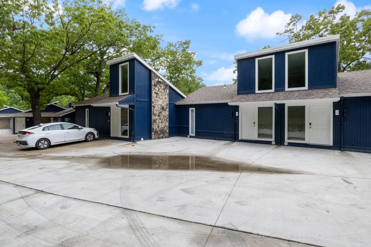 A modern condo exterior with blue siding and stone accents, set in a welcoming environment. The driveway is visible, along with lush greenery around the property. The open space in front of the home is ideal for outdoor activities, social gatherings, or letting pets enjoy the outdoors.