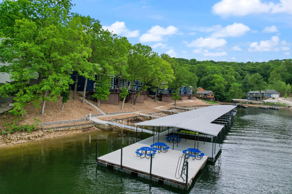 The image showcases a peaceful lakeside dock furnished with inviting blue tables and chairs, surrounded by lush greenery and clear, calm water. The dock provides direct access for boating and fishing, ideal for morning coffee, sunset views, and relaxed gatherings by the water. Nearby buildings indicate a friendly lakeside community while maintaining a sense of seclusion and tranquility.