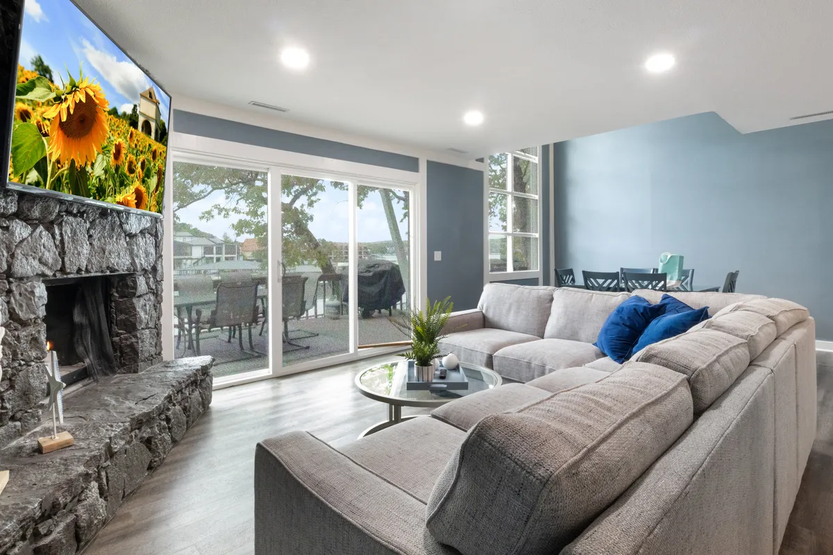 A large sectional sofa anchors the lounge area, which also boasts a stone fireplace, big windows with scenic views, a dining area visible in the background, and a large television. The space is comfortable and casual—ideal for both family movie nights and peaceful relaxation.
