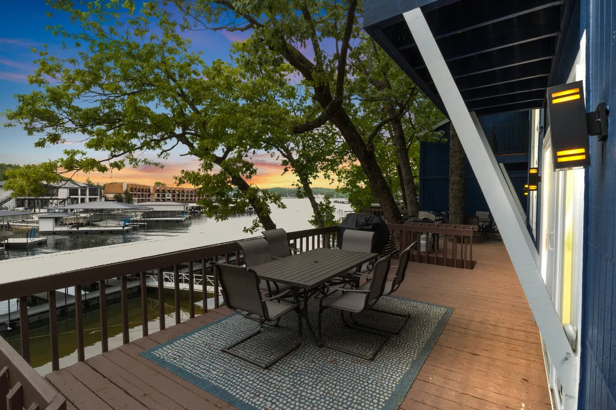 A broad deck with a dining table overlooks the shimmering lake, surrounded by trees and protected by a railing. The scene is tranquil, suggesting a space for outdoor dining, relaxation, and gathering while overlooking the water.