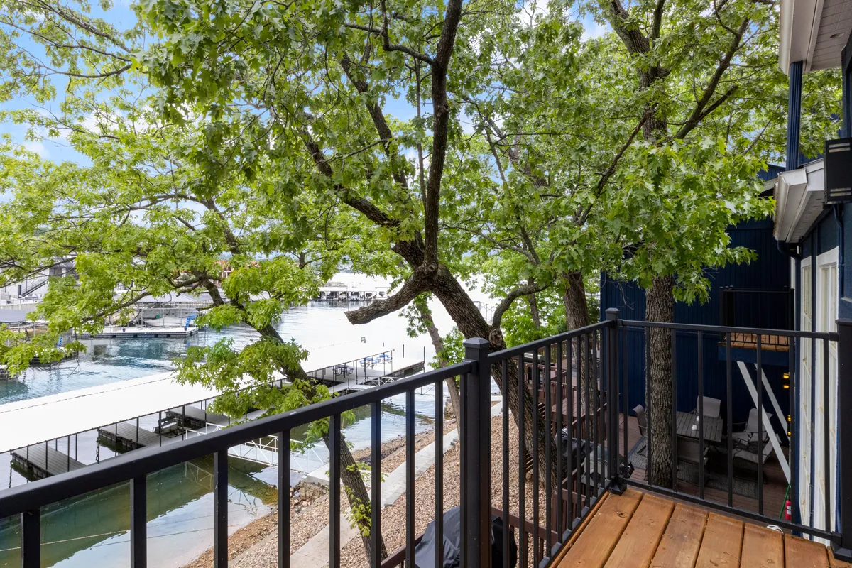 A serene waterfront view framed by lush green trees, as seen from a wooden balcony railing. The calm lake stretches into the distance, inviting quiet reflection or leisurely coffee moments while watching boats gently pass by.