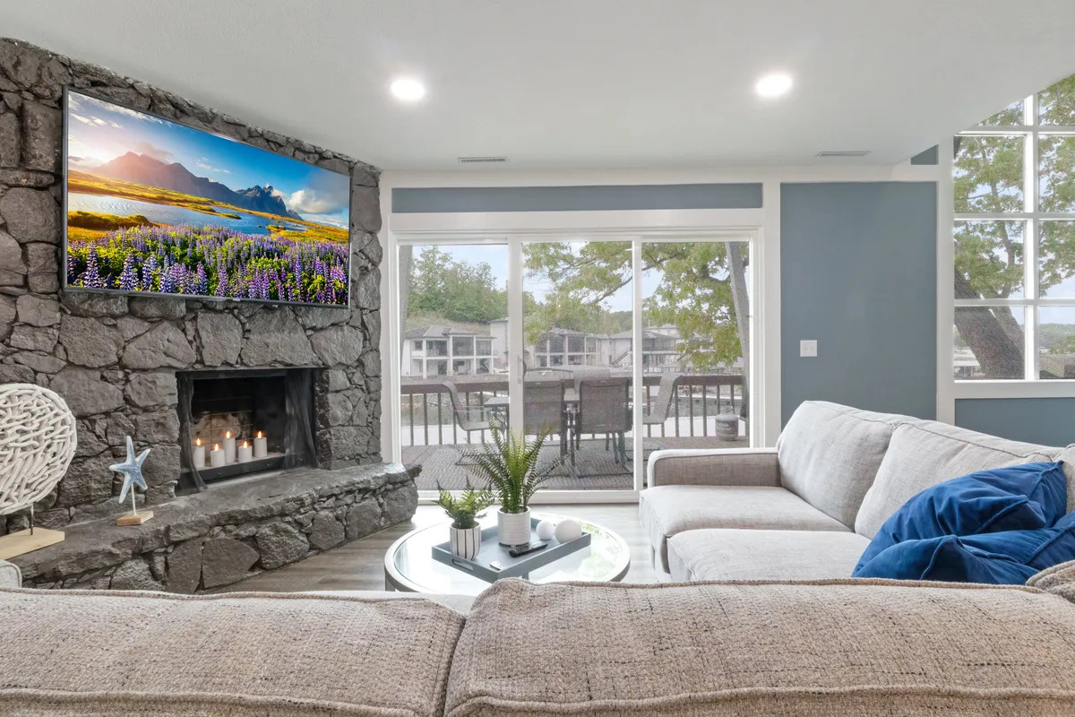A cozy and inviting lounge space with a stone fireplace, large television, comfortable sofa, a coffee table adorned with tasteful decor, and expansive windows framing serene outdoor views. The area is styled to foster both relaxation and entertaining.