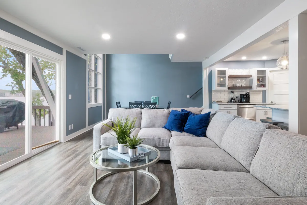 A spacious living room featuring a large grey sofa set against a blue accent wall, a glass coffee table, and expansive windows that allow natural light to flood the space. The open layout is visible, with comfortable seating inviting relaxation or social gatherings.