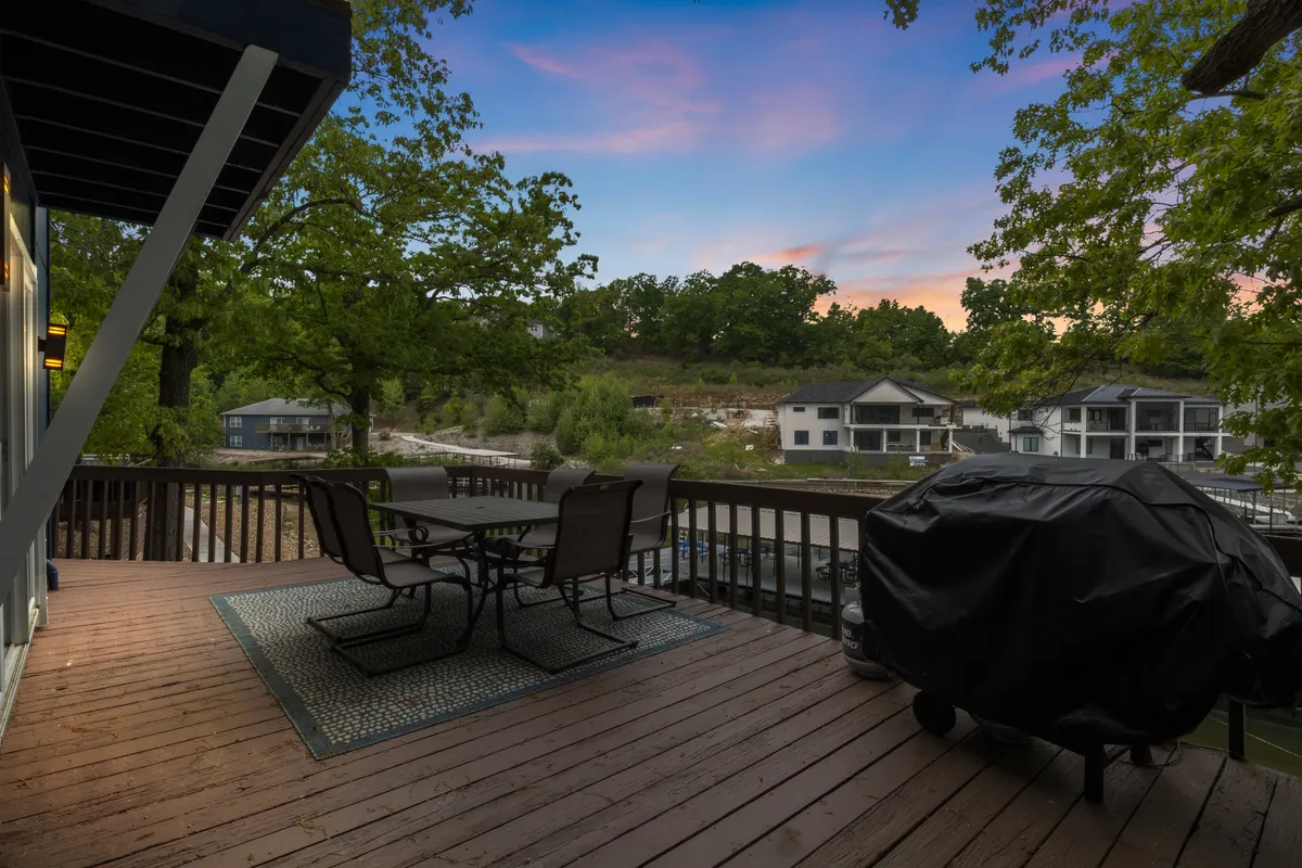 An inviting outdoor dining set and grill are set up on a patio overlooking the water. The sun casts a golden glow on the lake beyond, creating a picturesque setting for al fresco meals or lakeside gatherings. The space is ideal for group activities such as outdoor dining, evening drinks, or games by the water.