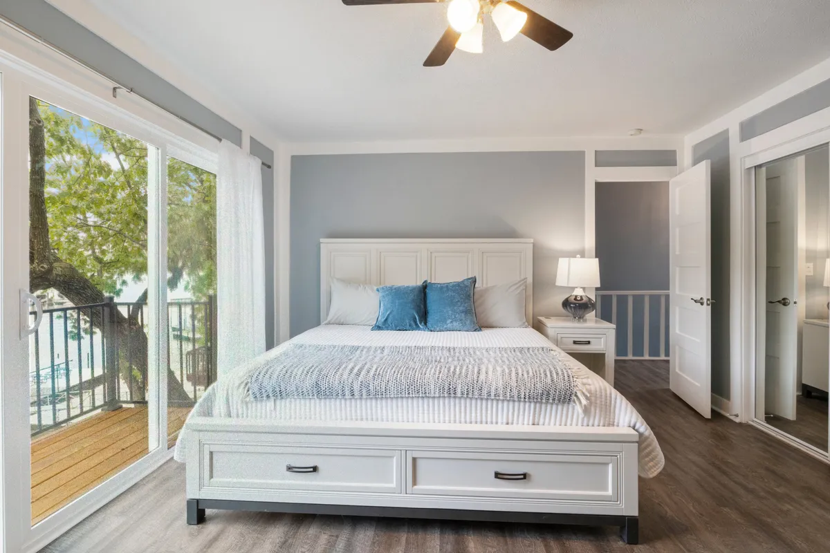 A cozy bedroom with a comfortable bed, light streaming in through wide windows, a nightstand with a lamp, and simple yet soothing decor. The space appears inviting and private, ideal for unwinding after a day of activity.