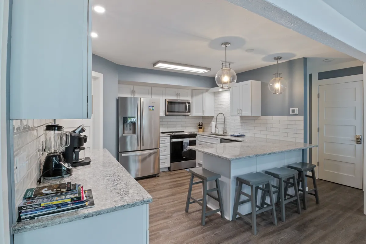 A bright, modern kitchen with a spacious island counter featuring stools, stainless steel appliances, granite countertops, and stylish hanging light fixtures. The layout invites interaction, with ample space for preparing meals and entertaining guests.